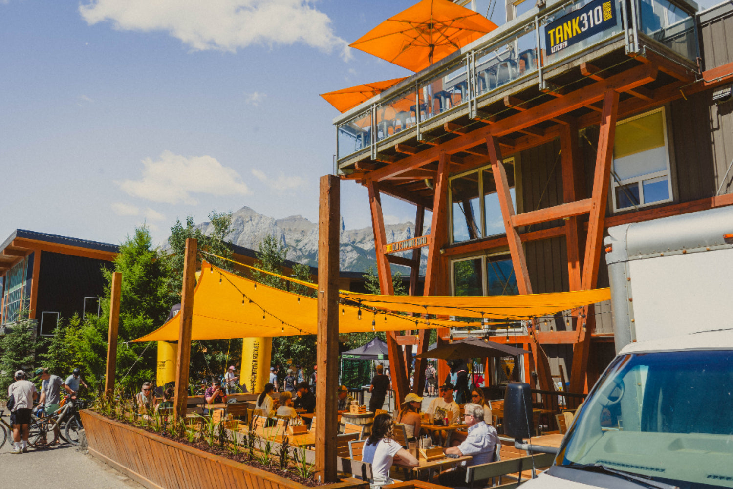 Outdoor dining area with wooden structure, orange umbrellas, and people sitting at tables under a blue sky.