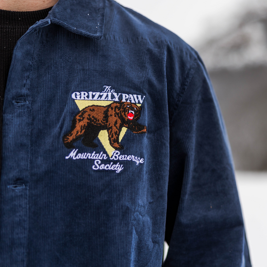 Navy blue jacket with Grizzly Paw Mountain Beverage Society logo on a blurred natural background