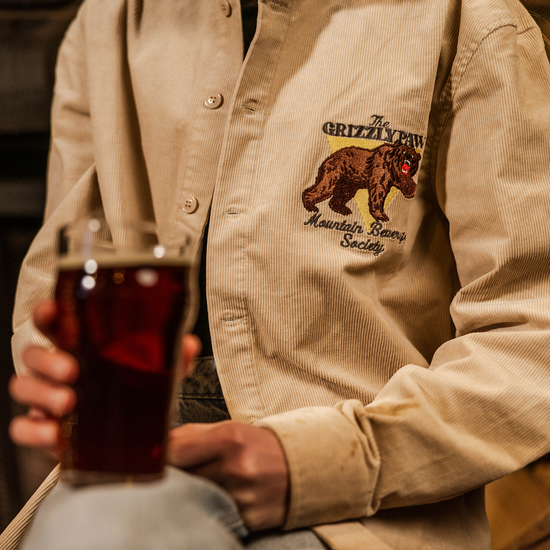 Person wearing a beige jacket with a bear graphic, holding a glass of dark liquid in front of a fireplace.