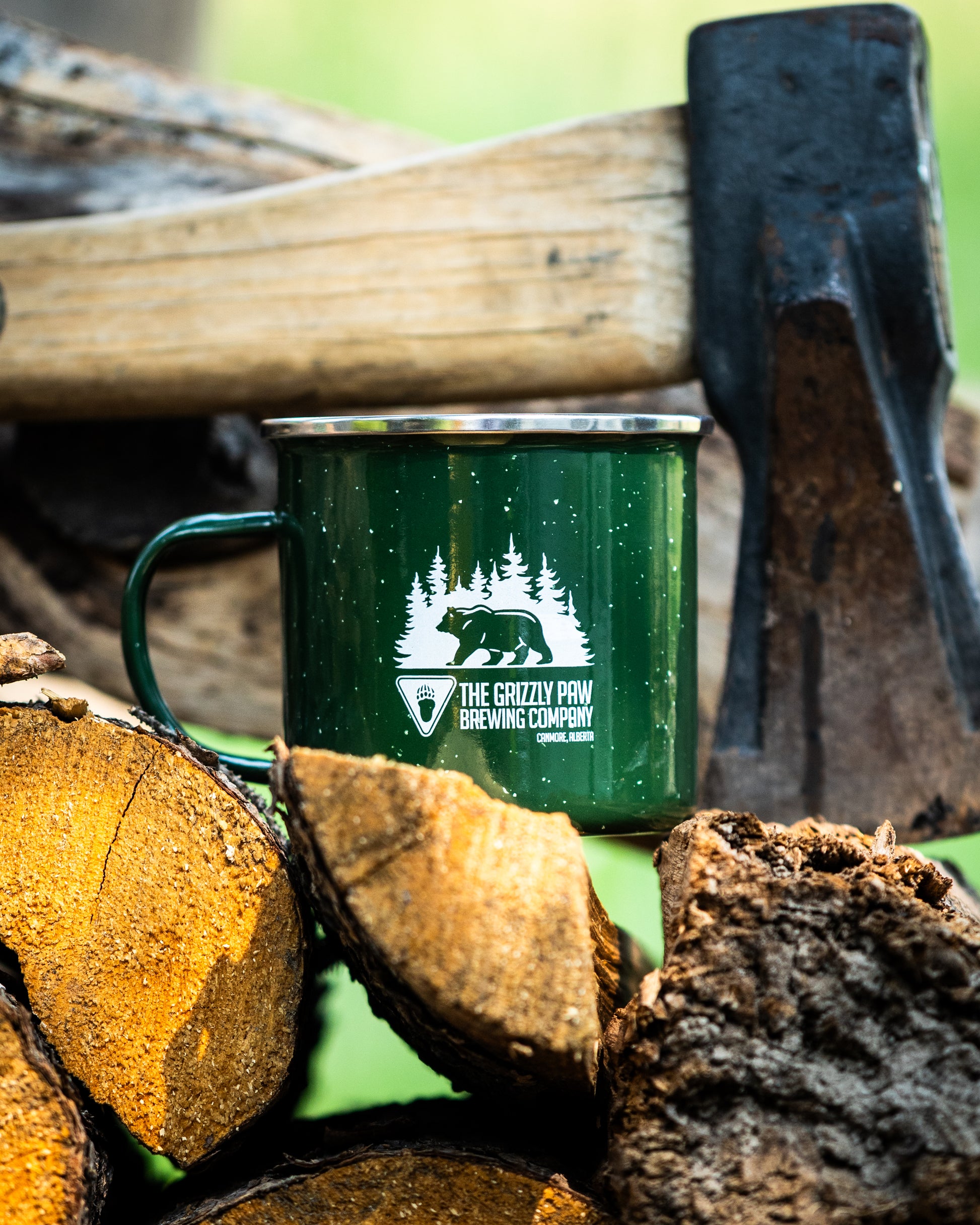 A dark green enamel camping mug with a white graphic of a bear and pine trees, resting on a pile of firewood with an axe in the background.