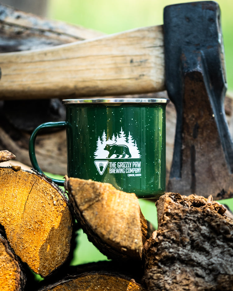 A dark green enamel camping mug with a white graphic of a bear and pine trees, resting on a pile of firewood with an axe in the background.