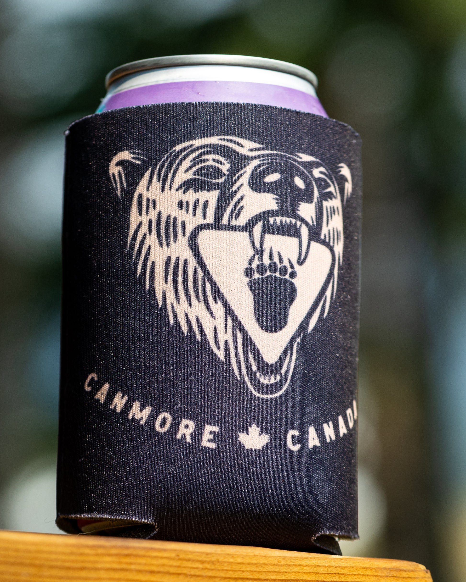 A black neoprene can cooler sleeve with a cream-colored grizzly bear graphic and the text 'CANMORE CANADA' sits on a wooden surface with a blurred outdoor background.