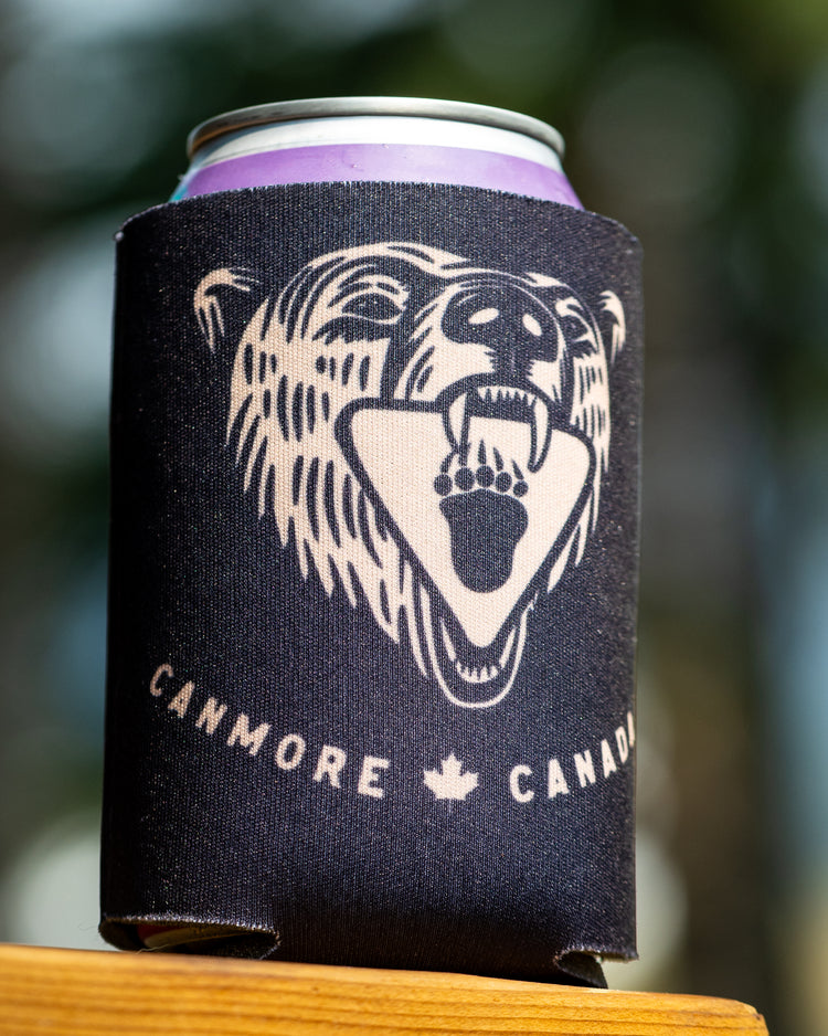 A black neoprene can cooler sleeve with a cream-colored grizzly bear graphic and the text 'CANMORE CANADA' sits on a wooden surface with a blurred outdoor background.