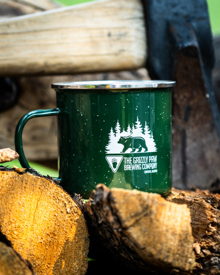 A dark green enamel camping mug with a white 'The Grizzly Paw Brewing Company' logo, resting on rustic logs outdoors.