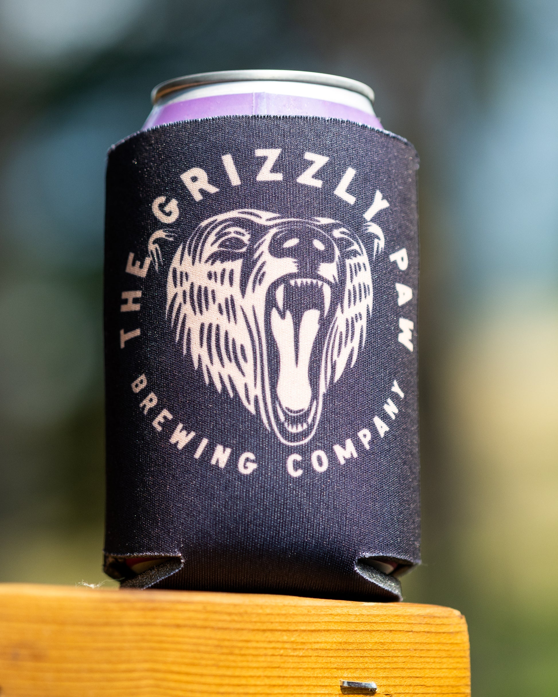 A black neoprene can cooler sleeve with a white roaring bear graphic and the text 'THE GRIZZLY PAW BREWING COMPANY' holding a purple-tinted beverage can, resting on a wooden surface with a blurred outdoor background.