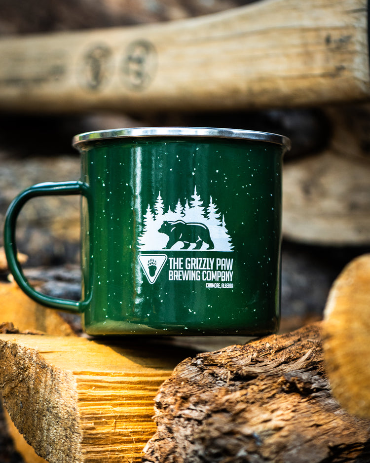 A dark green enamel camping mug with a white graphic of a bear and pine trees, featuring the text 'THE GRIZZLY PAW BREWING COMPANY', resting on a log with stacked wood logs in the background.