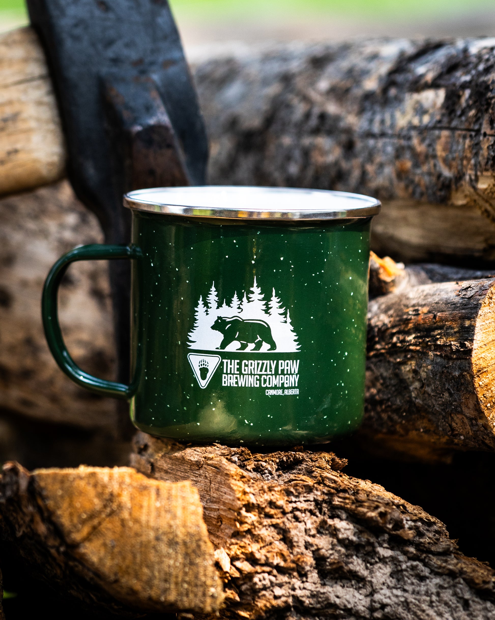A dark green enamel camping mug with a silver rim and handle, featuring a white graphic of a bear and pine trees with the text 'THE GRIZZLY POW BREWING COMPANY', set against a rustic background of stacked firewood logs.